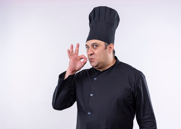 Männlicher Kochkoch, der schwarze Uniform und Kochhut trägt Zeichen für köstliches Stehen über weißem Hintergrund zeigt