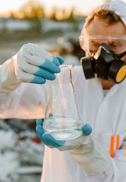 Männlicher Ökologe im Strahlenanzug, Gasmaske. Halten Sie das Reagenzglas mit Flüssigkeit, während Sie die Müllkippe studieren.