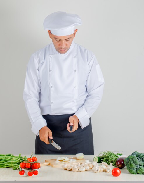 Männlicher Küchenchef, der Messer zum Hacken der Zwiebel in der Küche in Uniform, Hut und Schürze wählt