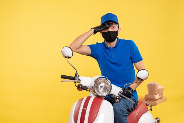 Männlicher Kurier der Vorderansicht, der auf dem Fahrrad in der Maske auf gelber Arbeitsservicid-einheitlicher Jobpandemielieferung sitzt