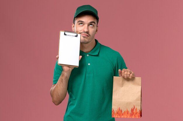 Männlicher Kurier der Vorderansicht im grünen einheitlichen Denken und Halten der Lebensmittelverpackung mit Notizblock auf hellrosa Hintergrund