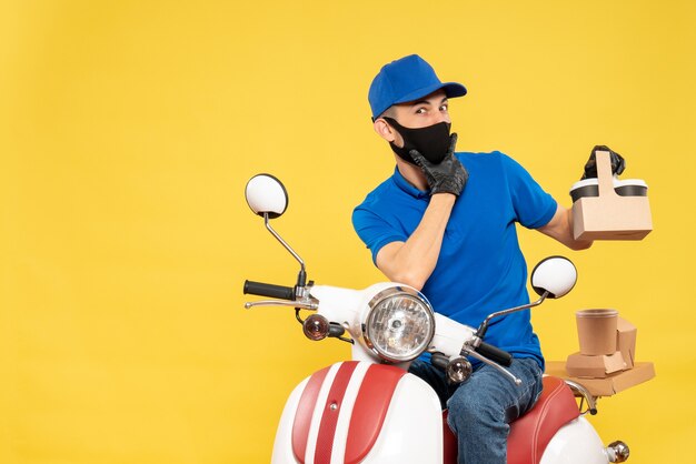 Männlicher Kurier der Vorderansicht in blauer Uniform mit Kaffee auf gelbem Pandemie-Fahrrad-Covid-Delivery-Virus-Arbeitsservice