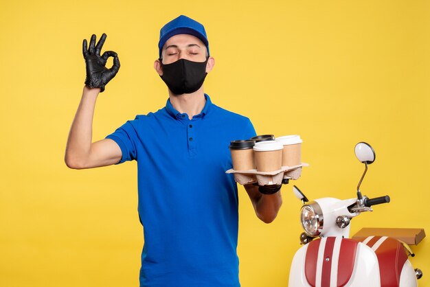 Männlicher Kurier der Vorderansicht in blauer Uniform und Maske, die Kaffee auf der gelben Uniform-Jobdienst-Covidwork-Lieferpandemie hält