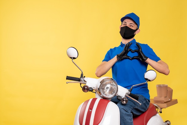 Männlicher Kurier der Vorderansicht in blauer Uniform und Maske, die Liebe auf gelbem Virusjobovid-Zustelldienstarbeitspandemie-Fahrrad senden