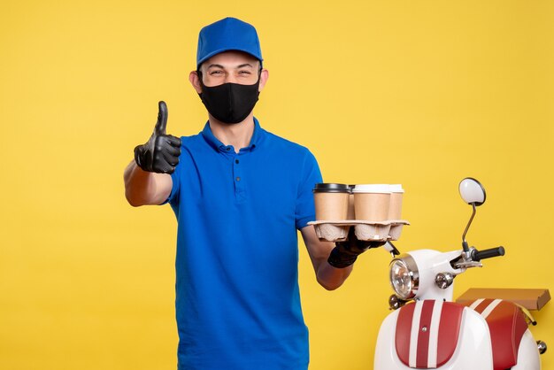 Männlicher Kurier der Vorderansicht in der blauen Uniform, die Kaffee hält und über gelbe Uniformfarbjob-Dienstkovidpandemie-Arbeitslieferung lacht