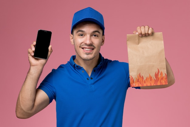 Männlicher kurier der vorderansicht in der blauen uniform, die lebensmittelpaket und telefon an der rosa wand hält, einheitliche servicebereitstellung