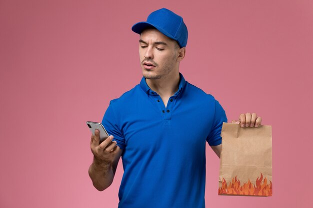 Männlicher Kurier der Vorderansicht in der blauen Uniform, die Lebensmittelpaket und Telefon auf rosa Wand hält, Jobuniform-Dienstlieferungsarbeiter