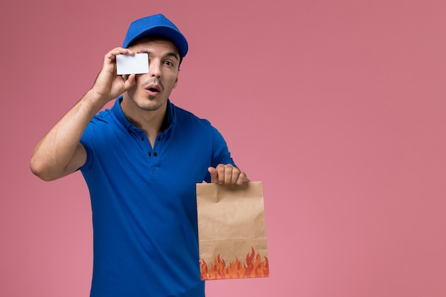 Männlicher Kurier der Vorderansicht in der blauen Uniform, die Lebensmittelverpackung mit Plastikkarte auf der rosa Wand hält, Arbeitsuniformuniformdienstlieferung