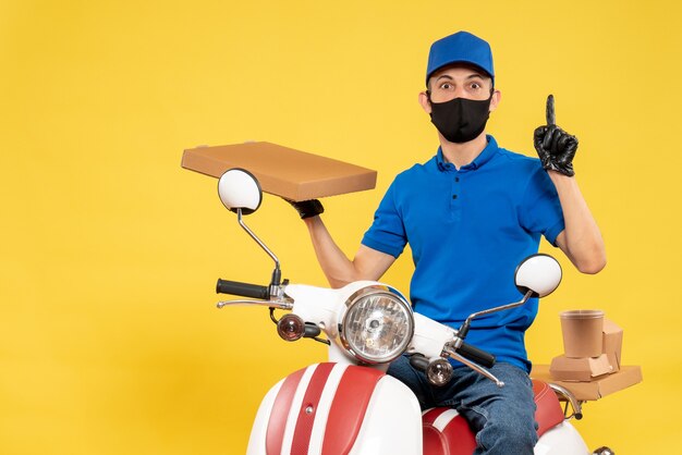 Männlicher Kurier der Vorderansicht in der blauen Uniform, die Nahrungsmittelbox auf gelbem Viruspandemie-Covid-Lieferarbeitsjob-Fahrrad hält