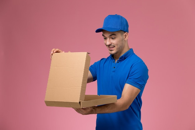 Männlicher kurier der vorderansicht in der blauen uniform, die öffnende nahrungsmittellieferbox an der rosa wand hält, einheitliche dienstauftragslieferung