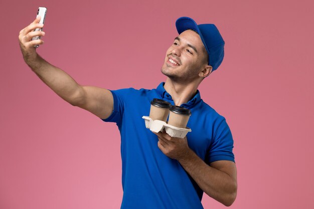Männlicher Kurier der Vorderansicht in der blauen Uniform, die selfie mit Kaffeetassen an der rosa Wand nimmt, einheitliche Dienstauftragszustellung