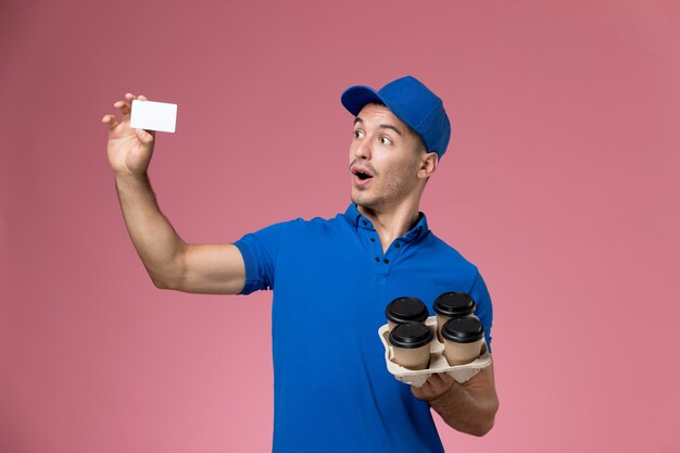 Männlicher Kurier der Vorderansicht in der blauen Uniform, die weiße Karte und Lieferkaffeetassen auf rosa Wand hält, einheitliche Jobarbeiterdienstlieferung