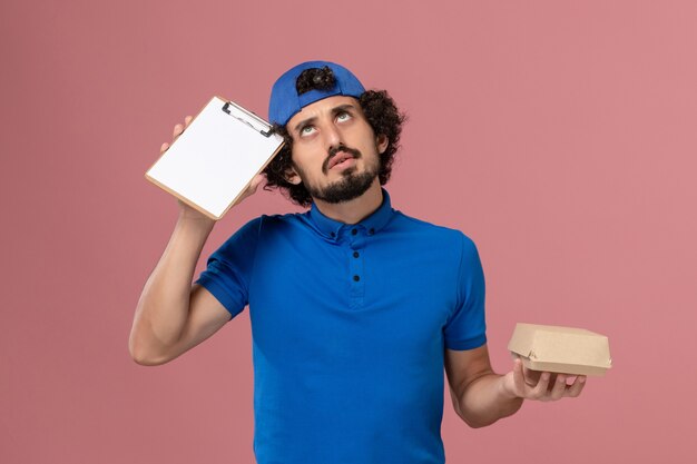 Männlicher Kurier der Vorderansicht in der blauen Uniform und im Umhang, die kleine Lieferung Lebensmittelpaket und Notizblock auf rosa Wanduniform-Lieferservice männlich halten