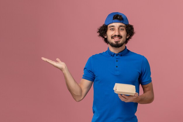 Männlicher Kurier der Vorderansicht in der blauen Uniform und im Umhang, die kleines Liefernahrungsmittelpaket an der rosa Wand halten