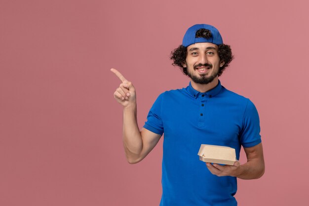 Männlicher Kurier der Vorderansicht in der blauen Uniform und im Umhang, die kleines Liefernahrungsmittelpaket an der rosa Wand halten