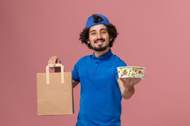 Männlicher Kurier der Vorderansicht in der blauen Uniform und im Umhang, die Lieferung Nahrungsmittelpaket und Schüssel auf dem rosa Wandlieferdienstuniformjob halten