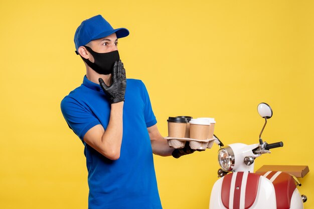 Männlicher Kurier der Vorderansicht in der blauen Uniform und in der Maske, die Kaffee auf gelber Arbeitsuniform-Jobdienst-Covid-Lieferpandemie hält