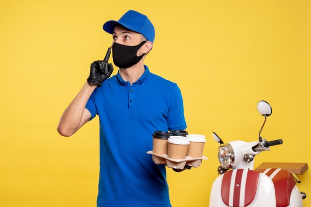 Männlicher Kurier der Vorderansicht in der blauen Uniform und in der Maske, die Kaffee auf gelber Arbeitsuniform-Jobdienst-Covid-Lieferung hält