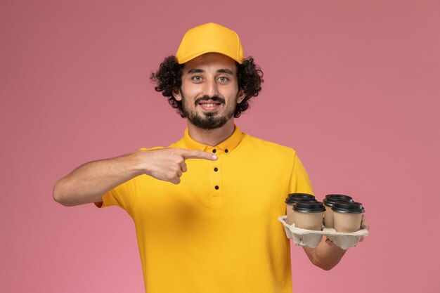 Männlicher Kurier der Vorderansicht in der gelben Uniform, die braune Kaffeetassen der Lieferung an der rosa Wand hält