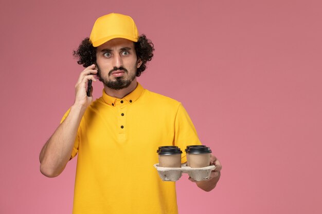 Männlicher Kurier der Vorderansicht in der gelben Uniform, die braune Kaffeetassen der Lieferung hält, die am Telefon auf rosa Wand sprechen