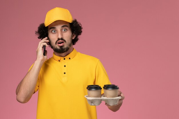 Männlicher Kurier der Vorderansicht in der gelben Uniform, die braune Kaffeetassen der Lieferung hält, die am Telefon auf rosa Wand sprechen