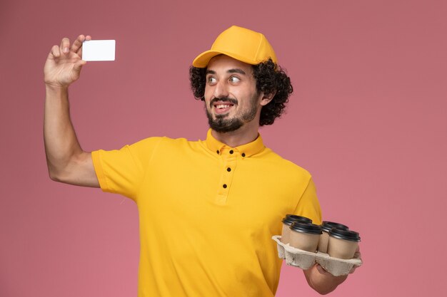 Männlicher Kurier der Vorderansicht in der gelben Uniform, die braune Kaffeetassen der Lieferung und weiße Karte an der rosa Wand hält