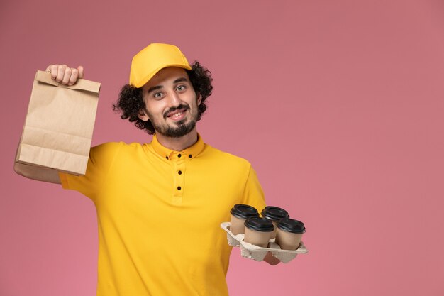 Männlicher Kurier der Vorderansicht in der gelben Uniform, die braune Kaffeetassen und Lebensmittelverpackung der Lieferung an der rosa Wand hält