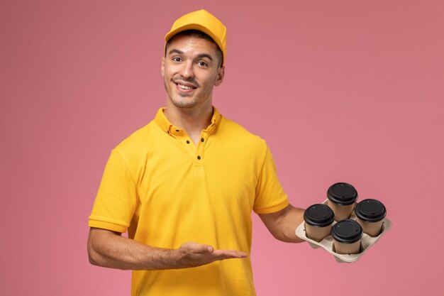 Männlicher Kurier der Vorderansicht in der gelben Uniform, die die Kaffeetassen der Lieferung auf rosa Hintergrund lächelt und hält