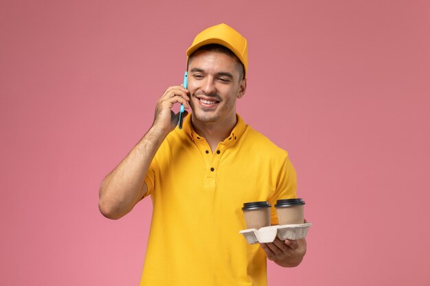 Männlicher Kurier der Vorderansicht in der gelben Uniform, die die Kaffeetassen der Lieferung hält, die am Telefon auf rosa Schreibtisch sprechen