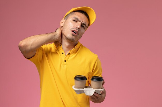 Männlicher Kurier der Vorderansicht in der gelben Uniform, die die Kaffeetassen der Lieferung hält, die Nackenschmerzen auf rosa Hintergrund haben