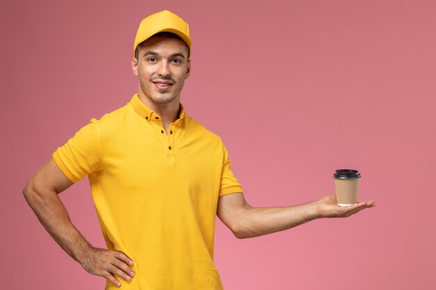 Männlicher Kurier der Vorderansicht in der gelben Uniform, die Kaffee-Lieferbecher auf dem rosa Schreibtisch hält