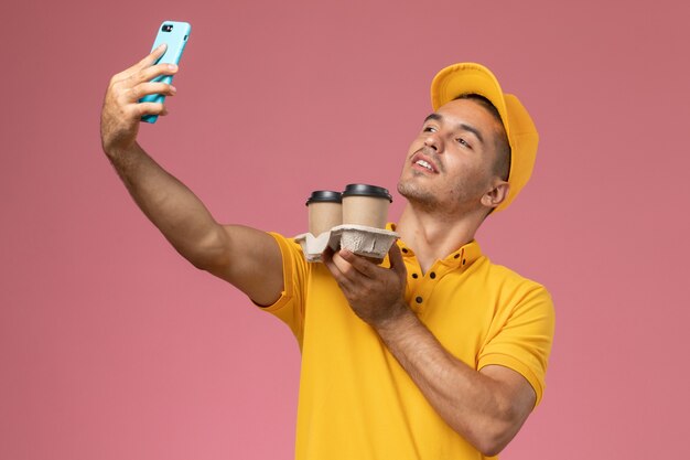 Männlicher Kurier der Vorderansicht in der gelben Uniform, die Kaffeetassen der Lieferung hält und ein Selfie auf rosa Hintergrund nimmt