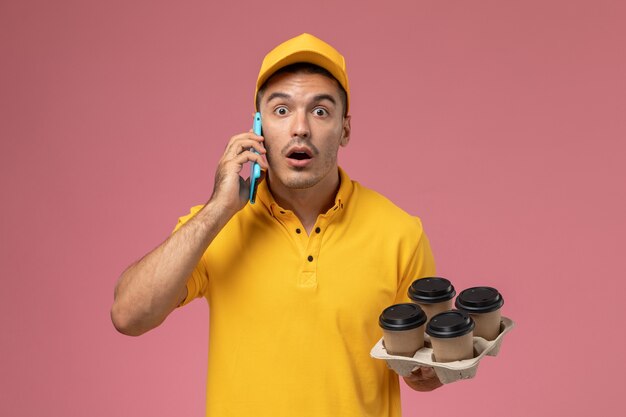 Männlicher Kurier der Vorderansicht in der gelben Uniform, die Kaffeetassen hält und am Telefon überrascht auf hellrosa Schreibtisch spricht