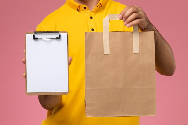 Männlicher Kurier der Vorderansicht in der gelben Uniform, die kleinen Notizblock und Liefernahrungsmittelpaket auf dem rosa Hintergrund hält.