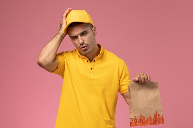 Männlicher Kurier der Vorderansicht in der gelben Uniform, die Kopfschmerzen hat und Nahrungsmittelpaket auf dem rosa Hintergrund hält