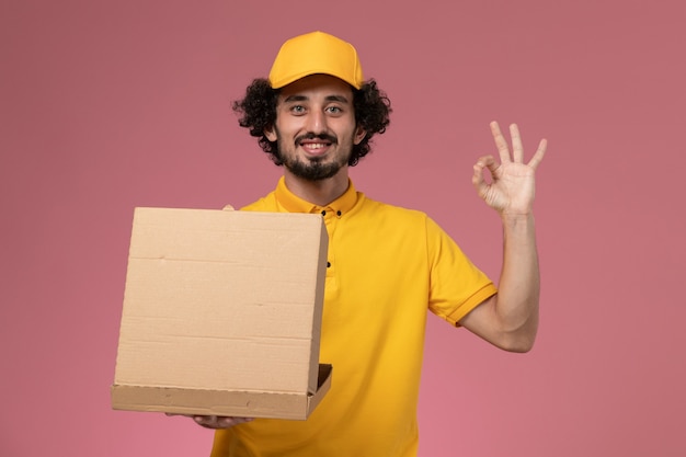 Männlicher Kurier der Vorderansicht in der gelben Uniform, die Lebensmittelabgabebox an der hellrosa Wand hält