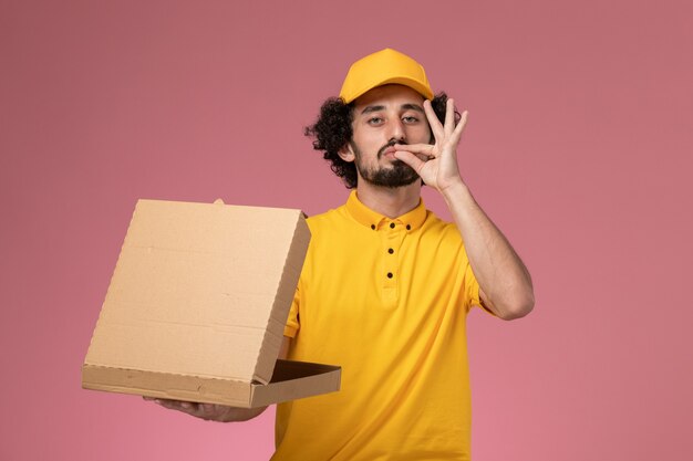 Männlicher Kurier der Vorderansicht in der gelben Uniform, die Lebensmittelabgabebox an der hellrosa Wand hält