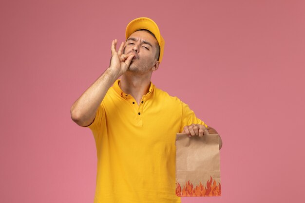 Männlicher Kurier der Vorderansicht in der gelben Uniform, die Lebensmittelpaket hält, das schmackhaftes Zeichen auf rosa Hintergrund zeigt