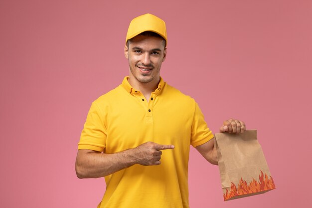 Männlicher Kurier der Vorderansicht in der gelben Uniform, die Lebensmittelpaket mit Lächeln auf rosa Hintergrund hält