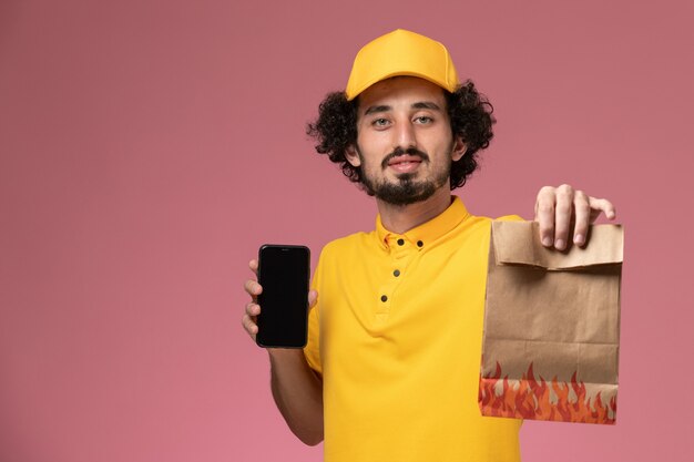 Männlicher Kurier der Vorderansicht in der gelben Uniform, die Lebensmittelpaket und Smartphone an der rosa Wand hält