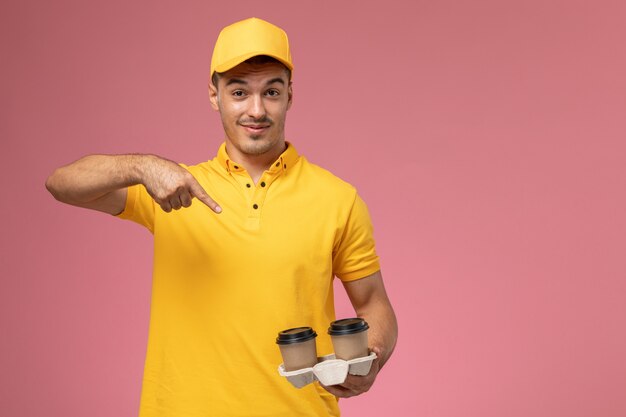 Männlicher Kurier der Vorderansicht in der gelben Uniform, die Lieferkaffeetassen auf hellrosa Schreibtisch hält