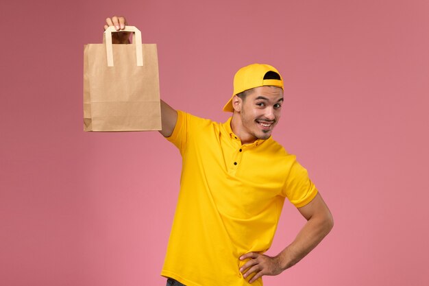 Männlicher Kurier der Vorderansicht in der gelben Uniform, die Lieferpapierpaket auf hellrosa Hintergrund hält.