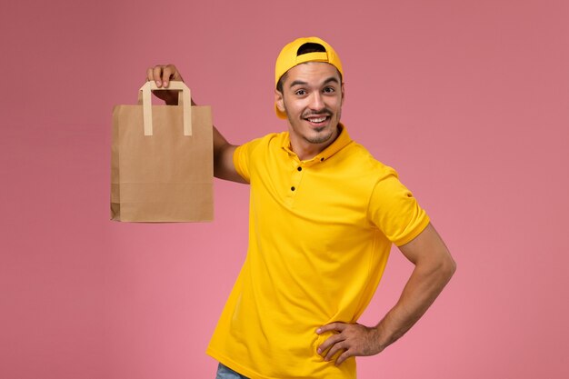 Männlicher Kurier der Vorderansicht in der gelben Uniform, die Lieferpapierpaket auf hellrosa Hintergrund hält.