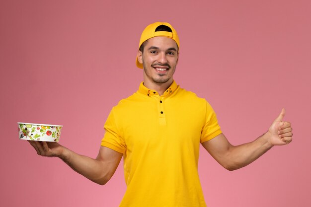 Männlicher Kurier der Vorderansicht in der gelben Uniform, die Lieferschalen hält, die auf dem rosa Hintergrund lächeln.