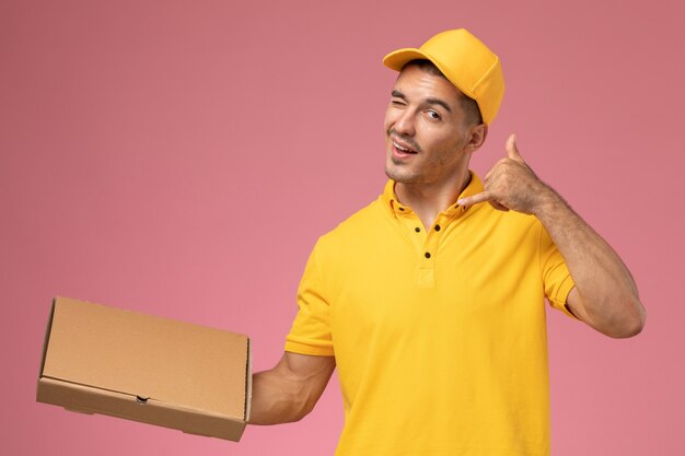 Männlicher Kurier der Vorderansicht in der gelben Uniform, die Nahrungsmittellieferbox auf rosa Schreibtisch hält
