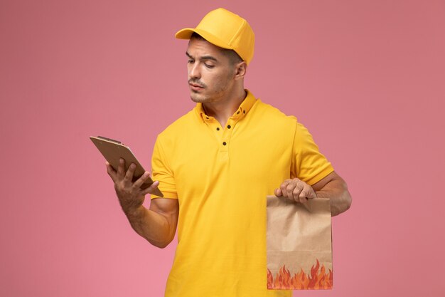 Männlicher Kurier der Vorderansicht in der gelben Uniform, die Notizblock liest und Nahrungsmittelpaket auf dem rosa Schreibtisch hält
