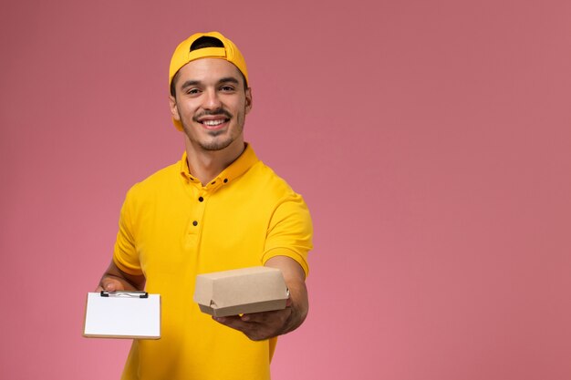 Männlicher Kurier der Vorderansicht in der gelben Uniform, die Notizblock und kleines Nahrungsmittelpaket auf rosa Hintergrund hält.