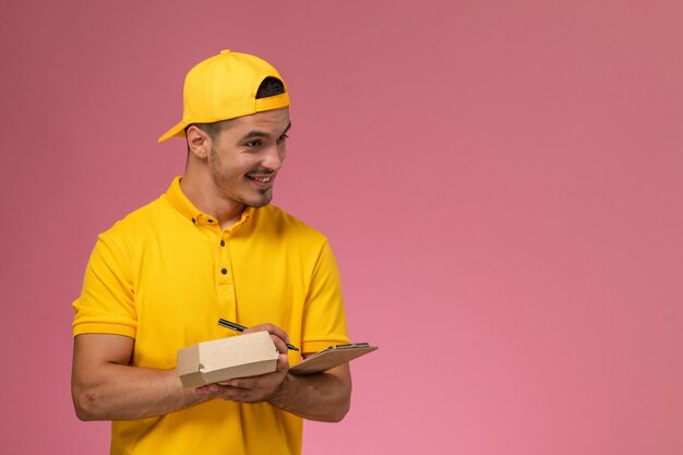 Männlicher Kurier der Vorderansicht in der gelben Uniform, die Notizblock und kleines Nahrungsmittelpaket hält und Notizen auf rosa Hintergrund schreibt.