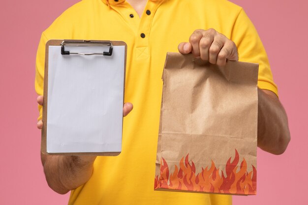 Männlicher Kurier der Vorderansicht in der gelben Uniform, die Notizblock und Nahrungsmittelpaket auf dem rosa Schreibtisch hält