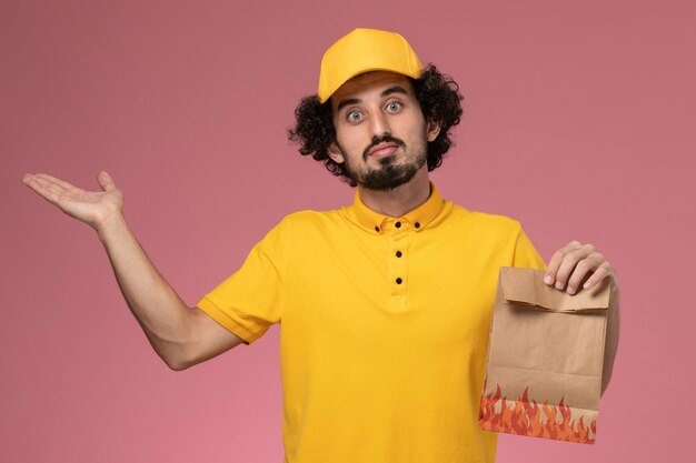 Männlicher Kurier der Vorderansicht in der gelben Uniform, die Papiernahrungsmittelpaket auf hellrosa Wand hält
